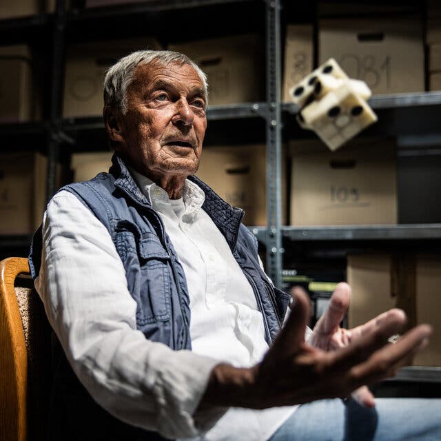 A portrait of Erno Rubik in a warehouse space, sitting in a chair next to table strewn with various Rubik’s-cube-like puzzles. He tosses one in his hand.