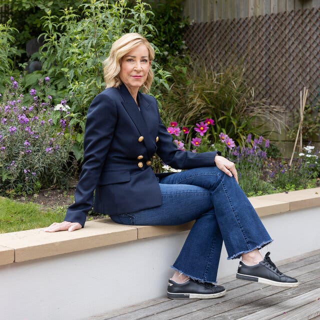 Chris Evert, the tennis champion, wearing a blue blazer with brass buttons, jeans and sneakers, sitting on a ledge in a garden.