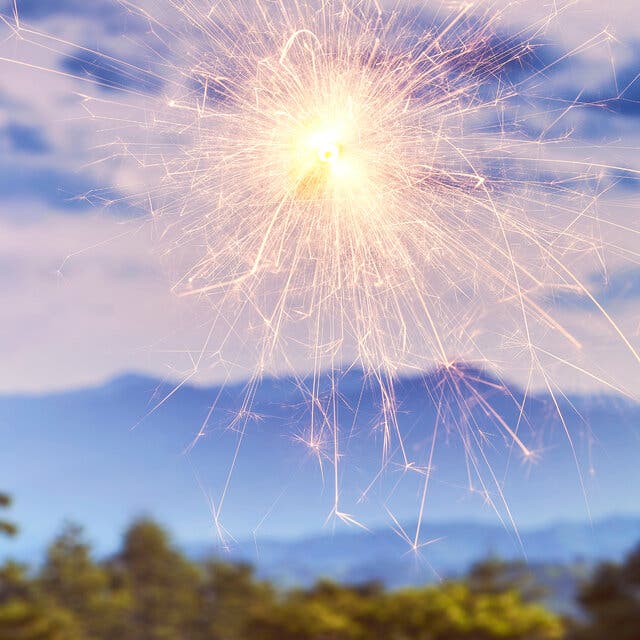 Strands of yellow light coming from a lit sparkler against a background of trees, mountains and sky. 