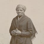 This sepia-toned photograph depicts a Black woman of middle age wearing a floor-length dark dress, a dark shawl and a head scarf. Her hands are clasped at her waist and she gazes directly into the camera.